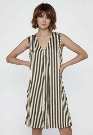 Giovane donna in piedi con le mani sui fianchi, che indossa un abito senza maniche al ginocchio con righe verticali bianche e nere su sfondo semplice.