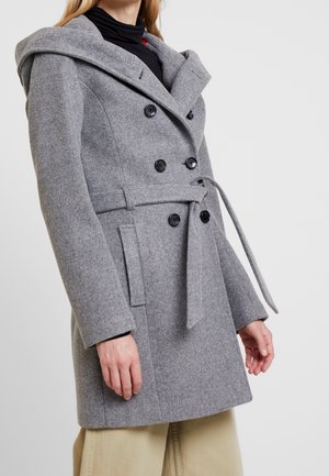 Kobieta w szarym, sięgającym do połowy długości płaszczu o dwurzędowym kroju, z paskiem i kapturem, w połączeniu z beżowymi spodniami i czarnym golfem.