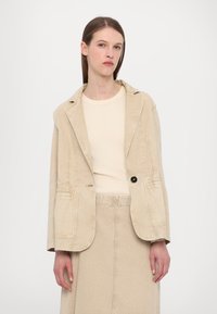 Beige corduroy bleiseri, jossa on yksi musta nappi, läpät, kaksi etutaskua ja rento istuvuus, yhdistettynä vaalean kermanväriseen puseroon.