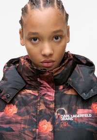 Casaco puffer com um padrão floral e de paisagens em tons escuros. Apresenta um colarinho alto, botões de pressão e um logótipo na área do peito.