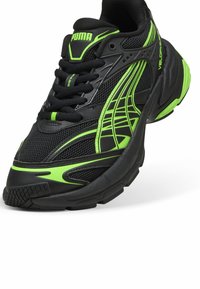 Zapatilla deportiva negra con detalles en verde, parte superior de malla, cierre de cordones y suela de goma texturizada. Presenta líneas de diseño elegantes y una marca identificativa.