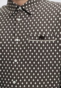 Gros plan d'une chemise foncée avec motif géométrique blanc, boutons blancs, poche devant avec logo brodé noir et col pointu.