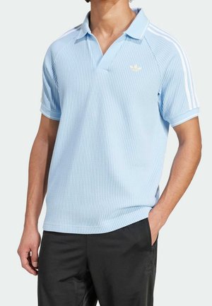 Homme portant un polo à manches courtes texturé bleu clair avec des rayures blanches sur les manches et un pantalon noir, main dans la poche, sur fond uni.