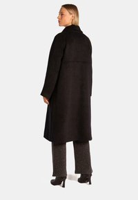 Lungo cappotto nero realizzato in tessuto morbido, con taglio dritto, ampio colletto e dettagli cuciti. Indossato sopra jeans scuri con decorazioni.