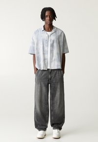 Chemise à manches courtes, boutonnée, avec un motif tie-dye bleu et blanc ; associée à un pantalon en denim gris surdimensionné et des baskets blanches.