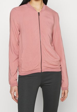 Femme portant une veste zippée rose clair à manches longues et un pantalon gris foncé sur un fond uni.