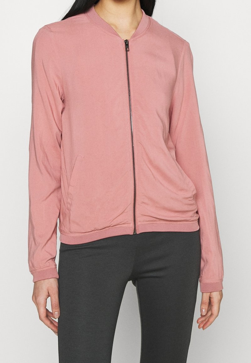 Femme portant une veste zippée rose clair à manches longues et un pantalon gris foncé sur un fond uni.