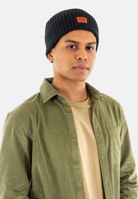 Marineblå ribbet hue med et mærke; grøn kordfløjls skjorte med knapper over en beige t-shirt; model som vender fremad.