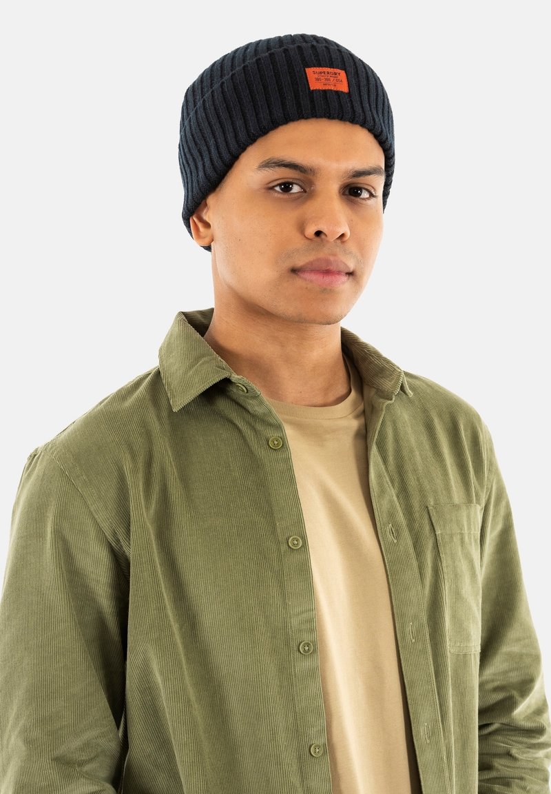 Marineblå ribbet hue med et mærke; grøn kordfløjls skjorte med knapper over en beige t-shirt; model som vender fremad.