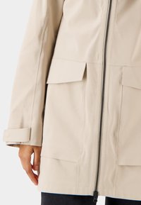 Béžová nepromokavá bunda s předním zipem, dvěma velkými klopovými kapsami a knoflíky na manžetách. Hladká textura a moderní design.