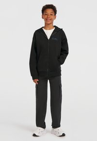 Čierna mikina na zips s prednými vreckami a modrým logom. Kombinovaná s čiernymi cargo nohavicami s bočnými vreckami a bielymi športovými topánkami.
