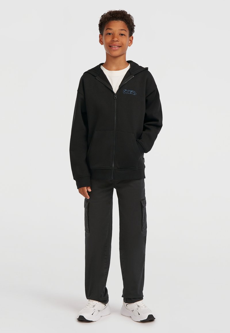 Čierna mikina na zips s prednými vreckami a modrým logom. Kombinovaná s čiernymi cargo nohavicami s bočnými vreckami a bielymi športovými topánkami.