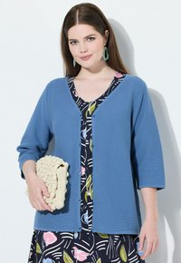 Světle modrý cardigan se třičtvrtinovými rukávy, zapínání na knoflíky, nošený přes vzorované šaty s květinovými a abstraktními vzory. Bílá háčkovaná taška.