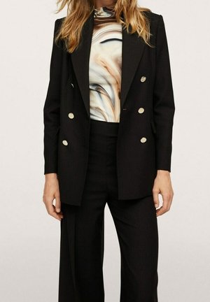 Mujer con blazer negro de doble botonadura con botones dorados, pantalones negros y una blusa de cuello alto con un patrón abstracto en beige y negro.
