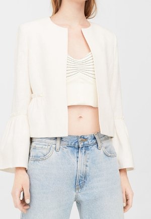 Kvinde iført en hvid cropped jakke med vide ærmer over en hvid ruched crop top og lyseblå højttaljede jeans.