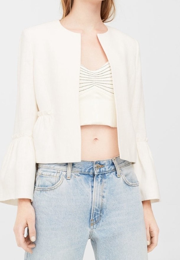 Femme portant une veste courte blanche à manches évasées sur un crop top blanc froncé et un jean taille haute bleu clair.
