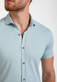 Šviesiai mėlynas polo marškinėliai su tekstūruotu audiniu, laisvas kirpimas, trumpomis rankovėmis, užsegamu apykaklės ir mažas logotipas kairiajame krūtinės šone.