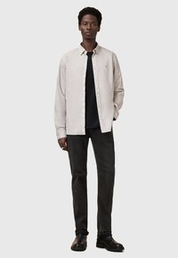 Lichtbeige knoopshirt over een zwart t-shirt met ronde hals, gecombineerd met donkergrijze jeans en zwarte laarzen; gladde stof, standaard kraag.