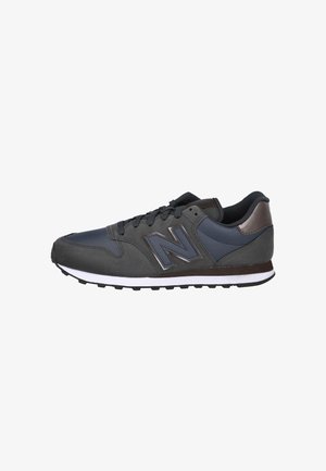 Zapatilla deportiva gris oscuro con material de ante y sintético, que presenta un acento en azul marino, una entresuela blanca y una suela exterior de goma negra.