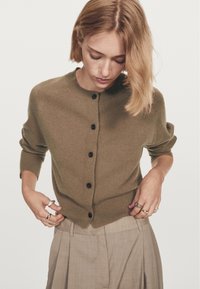 Olive green wool cardigan with a round neckline, six black buttons, and three-quarter sleeves, worn over light pleated trousers.