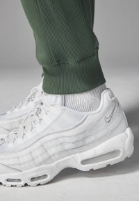 Białe sneakersy z teksturowanej siateczki i skórzanymi akcentami, z widoczną amortyzacją powietrzną. W połączeniu z zielonymi spodniami dresowymi z rowkami i szarymi skarpetkami.