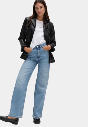 Kobieta w czarnej skórzanej marynarce, białej koszulce, jasnoniebieskich dżinsach o szerokich nogawkach i czarnych mokasynach, stojąca z rękami w kieszeniach.