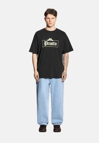 Czarna bawełniana koszulka z dużym białym logo "Prosto". Jasnoniebieskie dżinsy, luźny krój. Czarne buty z czerwonymi akcentami.