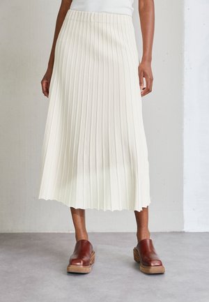 Woman wearing white pleated midi skirt with brown leather slip-on shoes, standing on gray floor against plain wall.