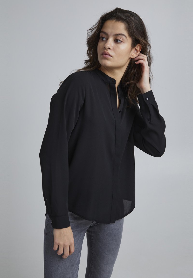 Zwarte blouse met lange mouwen en een mao-kraag, voorzien van een knopenlijst en manchetten, gecombineerd met grijze slimfit spijkerbroek.