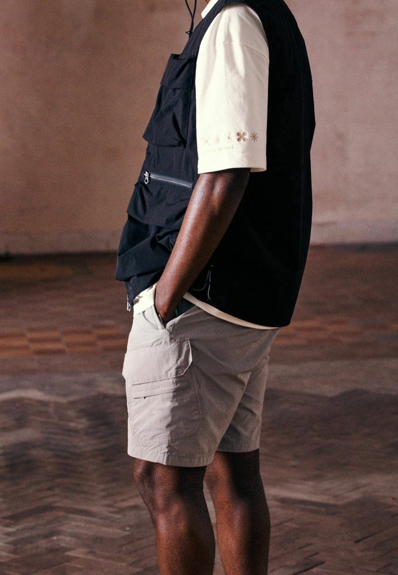 Personne portant un gilet noir avec plusieurs poches, une chemise crème à manches courtes et un short cargo beige, les mains dans les poches.