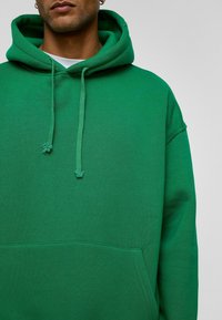 Groene hoodie van zacht materiaal met een voorzak, trekkoorden bij de capuchon en een relaxte pasvorm, waarbij de stiksels en textuur zichtbaar zijn.