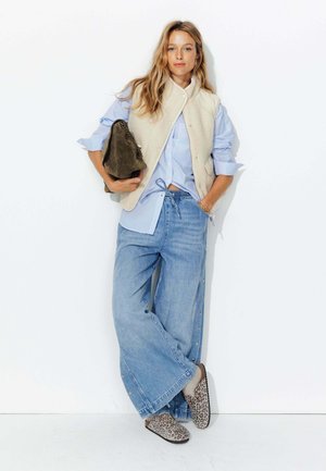 Gilet en polaire beige porté par-dessus une chemise à rayures bleu et blanc. Jeans larges bleu avec des boutons sur le côté. Chaussures à enfiler à motif animal.