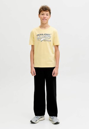 Camiseta de algodón amarilla con la inscripción "JACK & JONES Superior" en azul oscuro, combinada con pantalones negros de corte holgado y zapatillas grises.