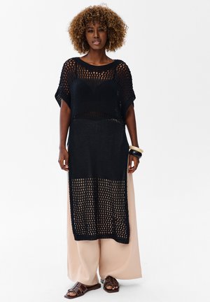 Kobieta o kręconych włosach, ubrana w czarną sukienkę z otwartym splotem na szerokich beżowych spodniach, brązowych sandałach i dużych bransoletkach na białym tle.