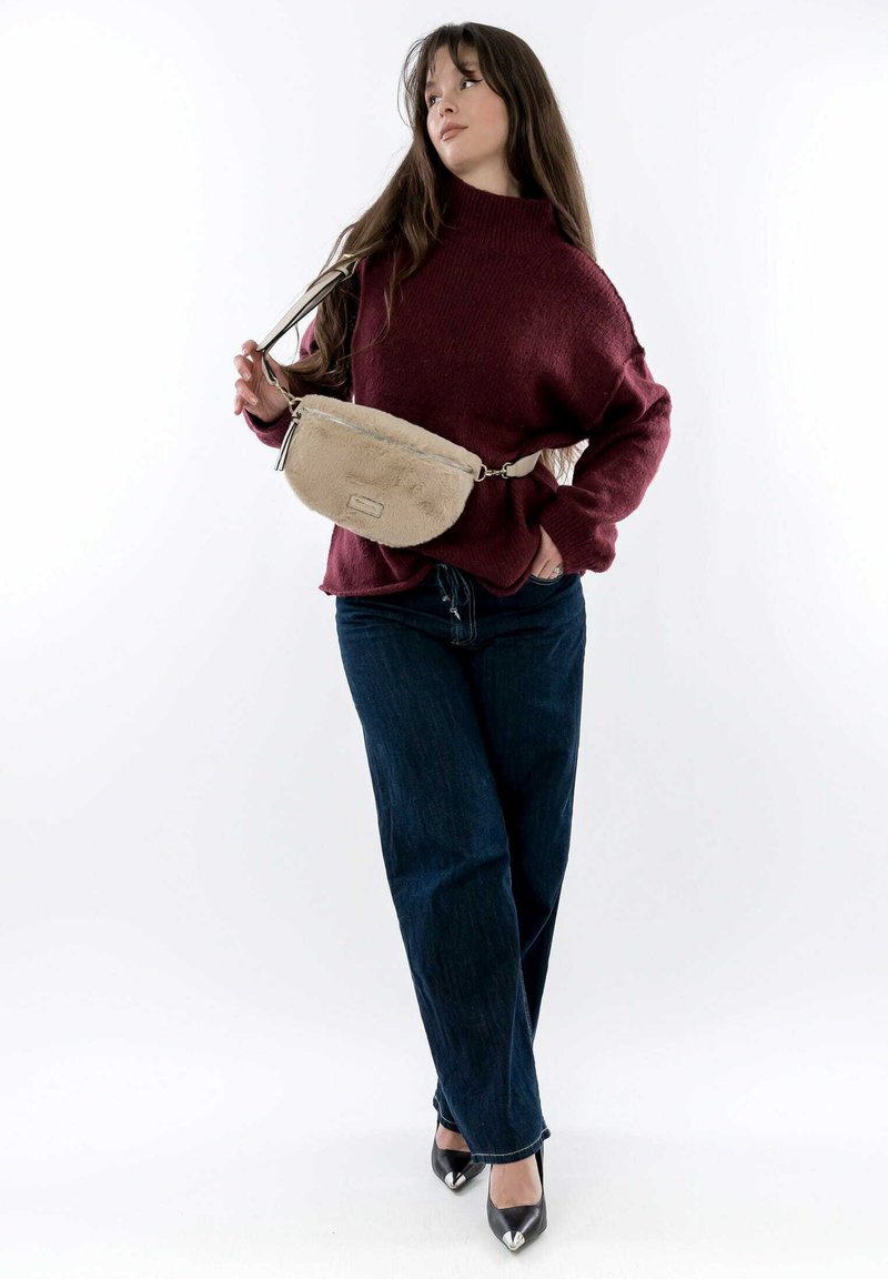 Pochette en fourrure beige avec une fermeture éclair argentée et une bandoulière, portée par-dessus un pull en tricot bordeaux, associée à un jean bleu foncé et des escarpins noirs.