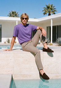 Jeune homme portant des lunettes de soleil, chemise violette, pantalon beige et chaussures marron, assis au bord de la piscine avec des palmiers et une maison moderne en arrière-plan.