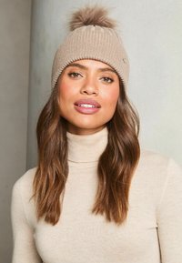 Beige knit beanie with a plush pom-pom on top. Features a ribbed texture and a small decorative badge on the front.