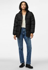 Svart puffjacka med huva, med kviltade sektioner och dragkedjestängning, bärs över en ljusfärgad topp och blåjeans.