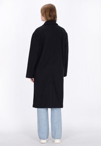 Cappotto lungo nero con colletto a punta e vestibilità oversize, realizzato in tessuto testurizzato. Abbinato a jeans a gamba larga blu chiaro e sneakers bianche.
