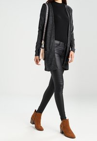 Szary, długi kardigan z kieszeniami noszony na czarnej dopasowanej bluzce, zestawiony z czarnymi skórzanymi legginsami i brązowymi botkami na masywnym obcasie.