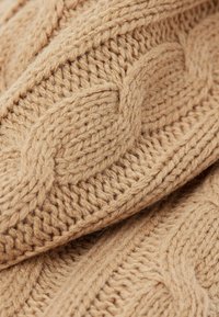 Strickpullover aus beigem Garn, der ein dickes Kabelmuster aufweist, mit sichtbarer Textur und verflochtenen Details im gesamten Stoff.