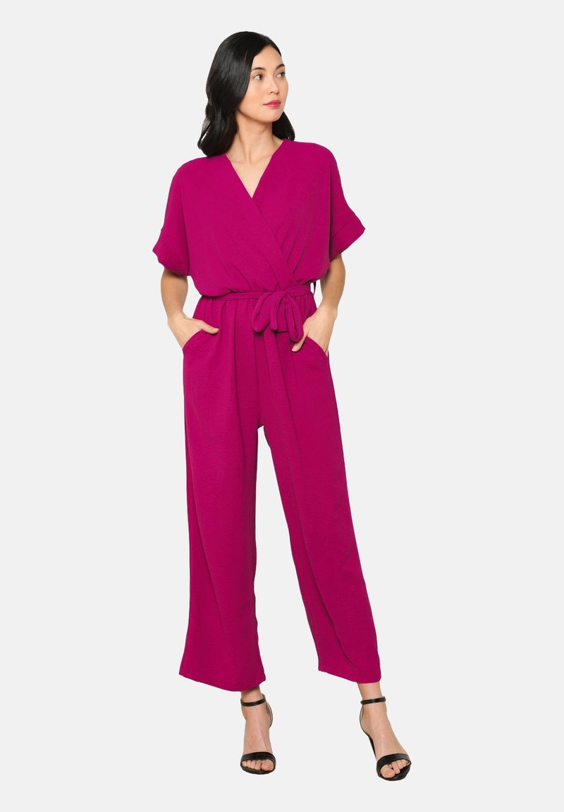 Fuchsia jumpsuit met een wijde broekspijp ontwerp, korte mouwen, gewikkelde voorkant en een gestrikt taille. Voorzien van twee zijzakken. Gdraagd met zwarte haksandalen.