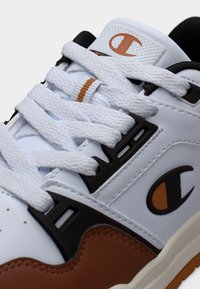 Λευκό αθλητικό παπούτσι με καφέ λεπτομέρειες, υφασμένους μαύρους επενδύσεις, παχιά λευκά κορδόνια και ένα σήμα με λογότυπο στη γλώσσα.