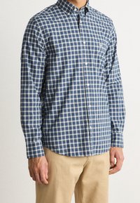 Niebiesko-biała koszula w kratę na guziki z długimi rękawami, kołnierzykiem oraz kieszenią na piersi. Materiał wydaje się lekki i gładki.