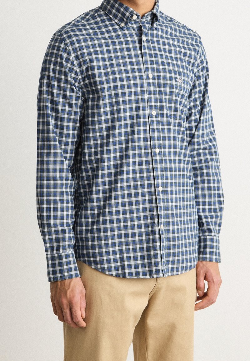 Niebiesko-biała koszula w kratę na guziki z długimi rękawami, kołnierzykiem oraz kieszenią na piersi. Materiał wydaje się lekki i gładki.