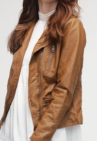 Brązowa skórzana kurtka o dopasowanym kroju, z asymetrycznym zamkiem z przodu, bocznymi kieszeniami na zamek i teksturowanym wykończeniem. Nosi się ją na białej sukience.