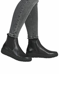 Piernas en jeans ajustados grises llevando botas de cuero negras hasta el tobillo con lados elásticos y cremallera lateral, de pie contra un fondo blanco.