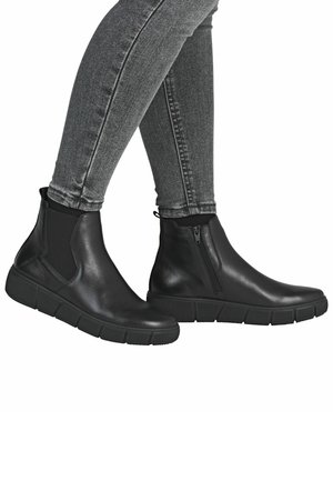 Jambes en jean skinny gris portant des bottes en cuir noir avec des côtés élastiques et une fermeture éclair sur le côté, debout contre un fond blanc.