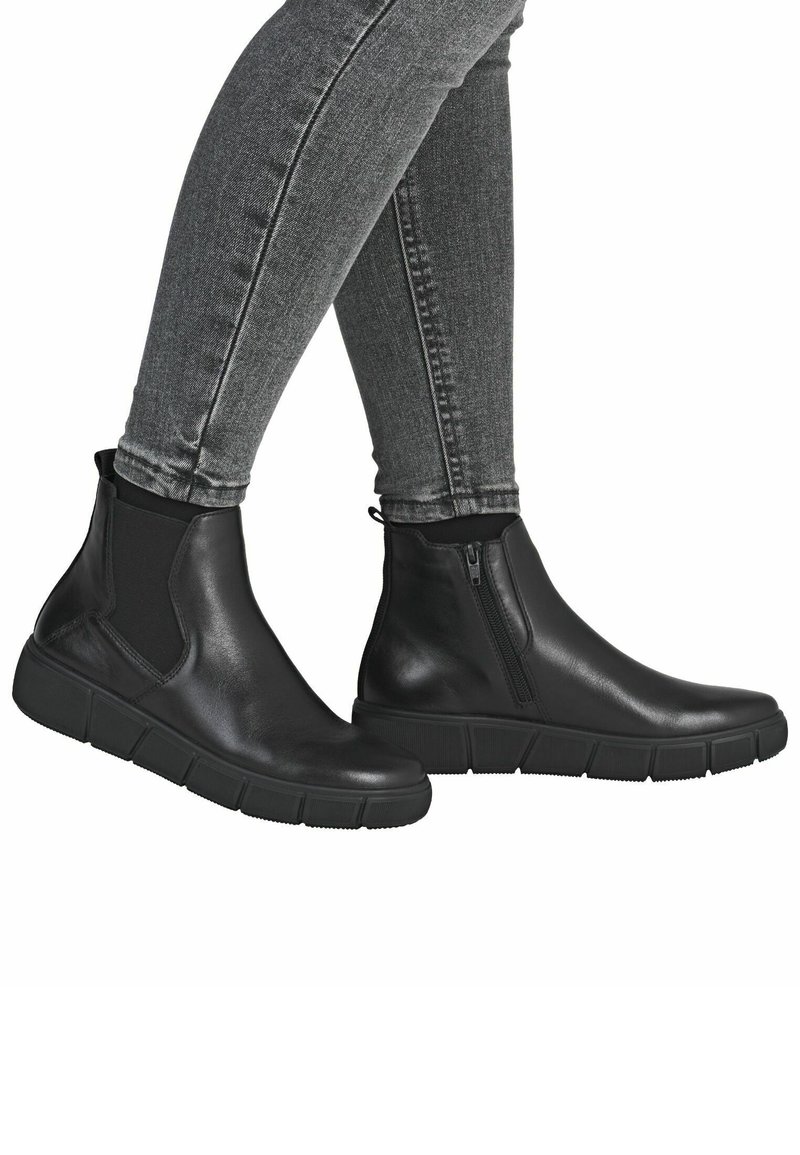 Piernas en jeans ajustados grises llevando botas de cuero negras hasta el tobillo con lados elásticos y cremallera lateral, de pie contra un fondo blanco.