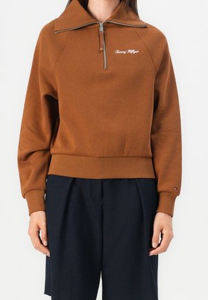 Femme portant un sweat-shirt marron à fermeture éclair au quart avec le logo "Tommy Hilfiger" et un pantalon large bleu marine, debout devant un fond uni.